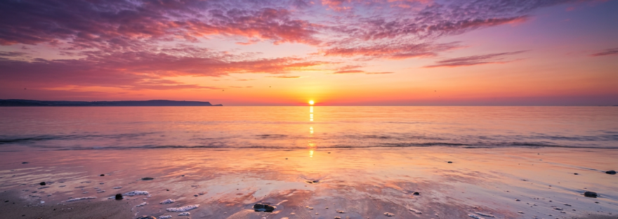 Sun setting over calm ocean with colorful sky and stones on sandy beach